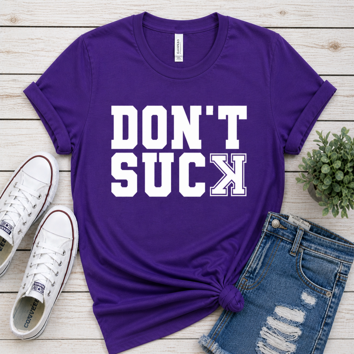 Purple t-shirt with 'DON'T SUCK' text on a wooden surface with shoes and jeans.