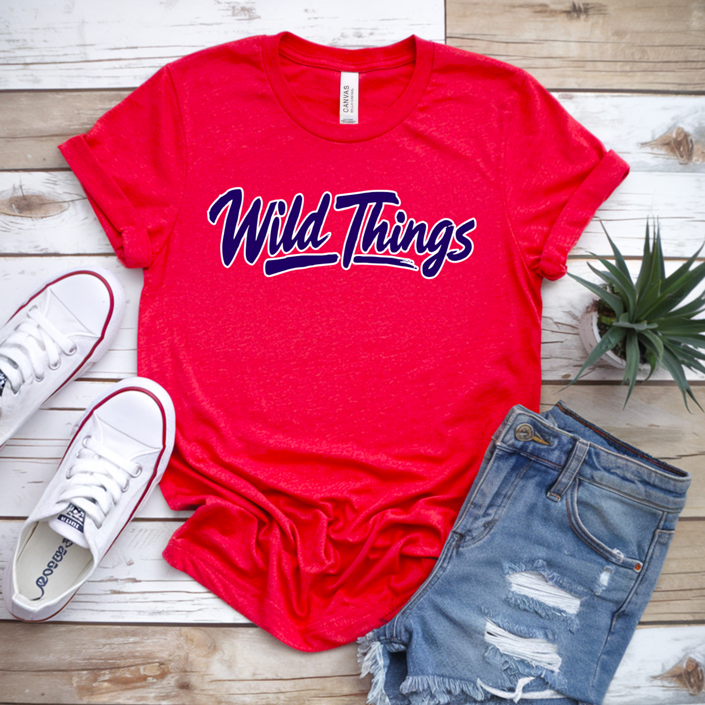 Red t-shirt with 'Wild Things' text, white sneakers, denim shorts, and a plant on a wooden surface.