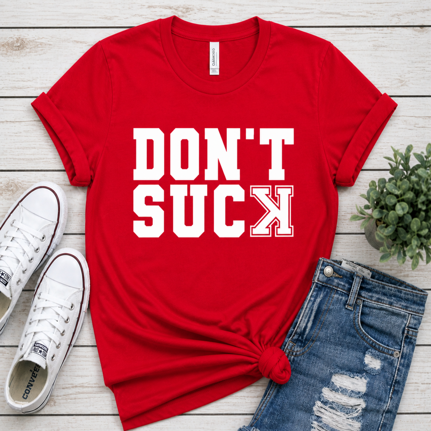 Red t-shirt with 'DON'T SUCK' text on a wooden surface with shoes and jeans.