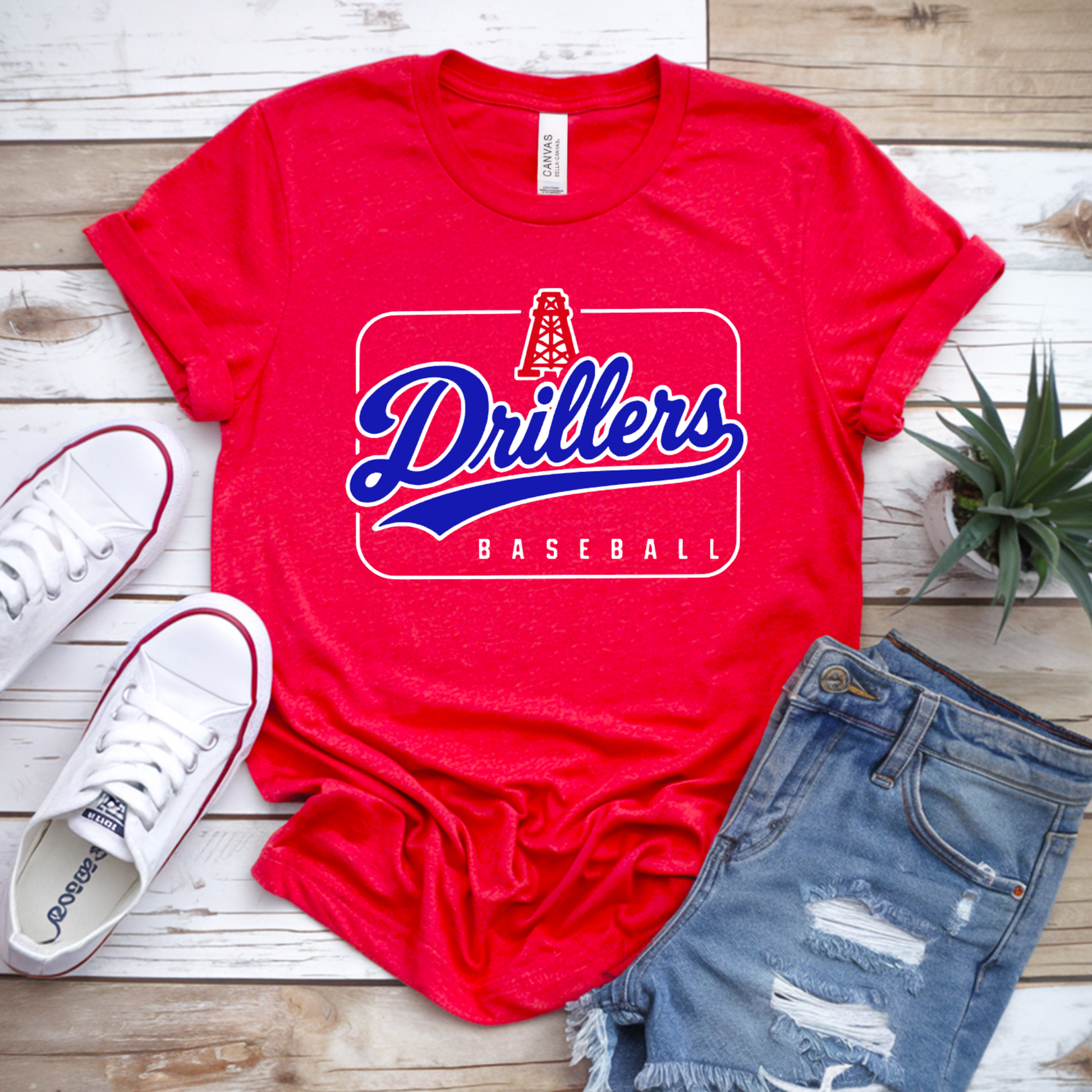 A red graphic T-shirt laid flat on a light wood surface, styled with white sneakers, distressed denim shorts, and a small potted plant. The shirt has a large front graphic reading "Drillers Baseball" in blue and white.