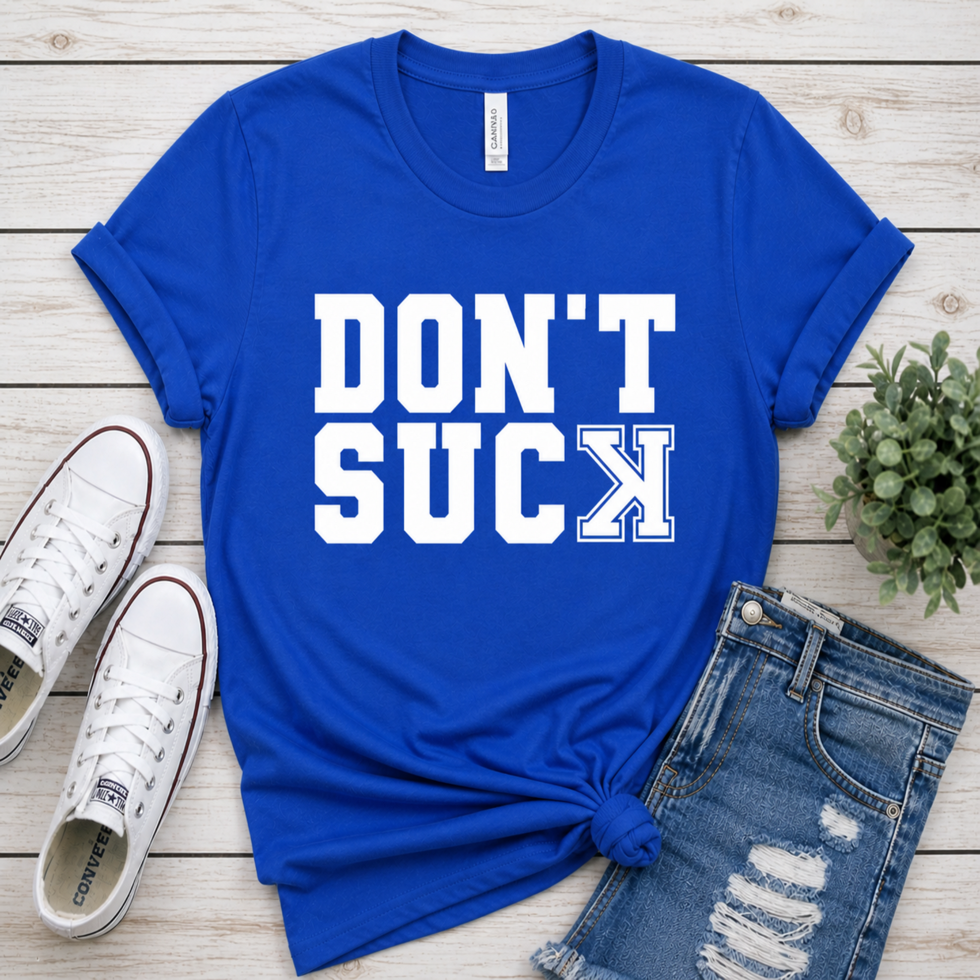Blue t-shirt with 'DON'T SUCK' text on a wooden surface with sneakers and jeans.