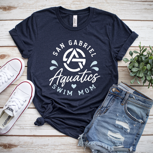 Navy blue t-shirt with 'San Gabriel Aquatics Swim Mom' design on a wooden surface with sneakers and shorts.