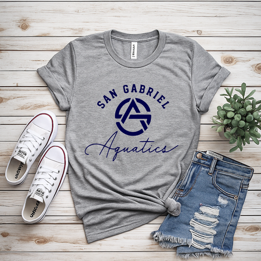 Gray t-shirt with 'San Gabriel Aquatics' logo, paired with sneakers and jeans on a wooden surface.