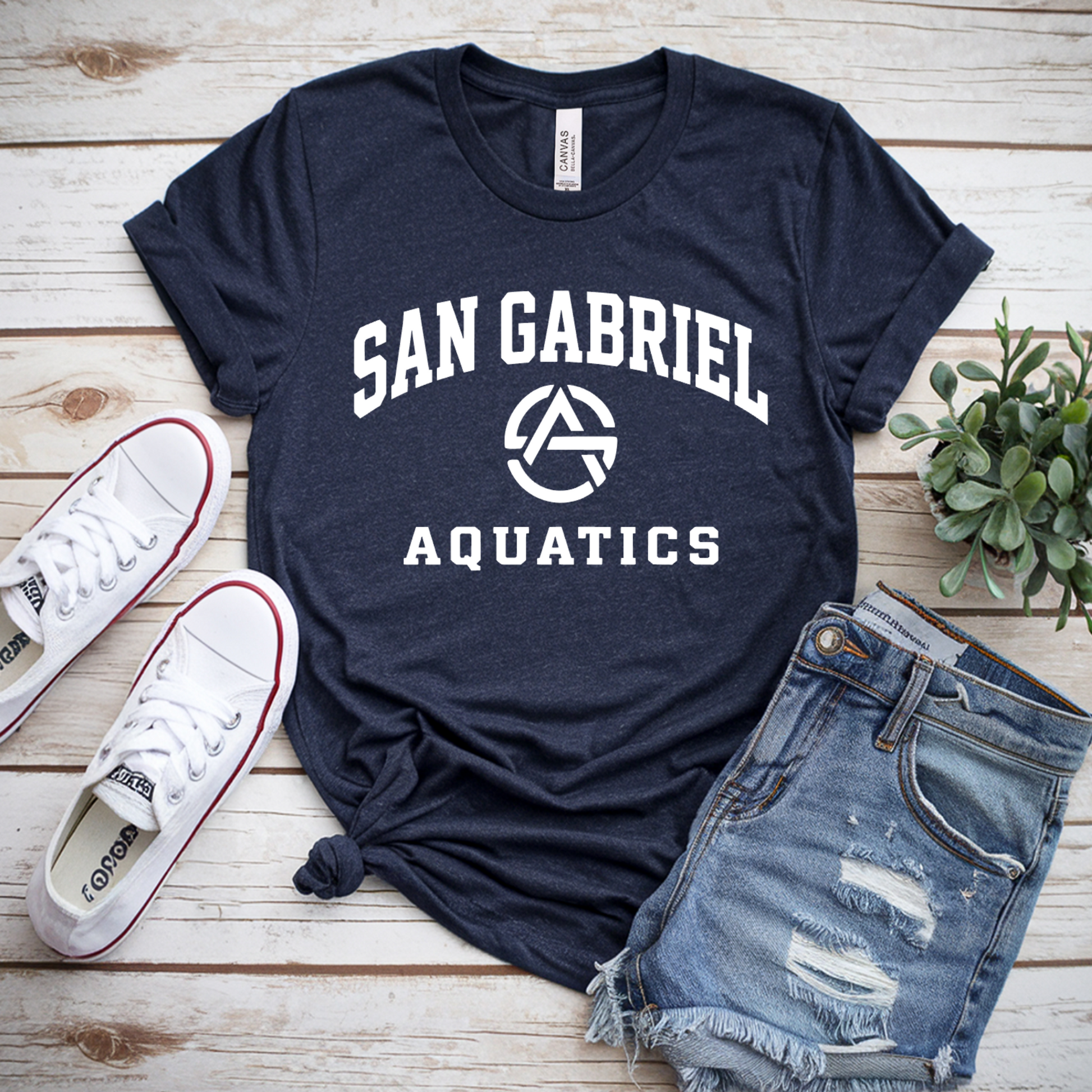 Navy blue t-shirt with 'San Gabriel Aquatics' text, white sneakers, and denim shorts on a wooden surface.