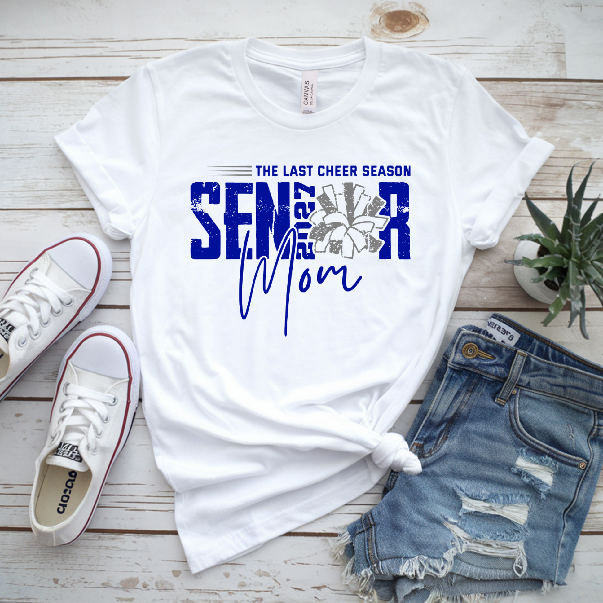 White graphic T-shirt laid flat on a whitewashed wood surface, styled with white sneakers, distressed denim shorts, and a small potted plant. The shirt reads 'THE LAST CHEER SEASON SENIOR Mom' in blue and gray text with a floral design.
