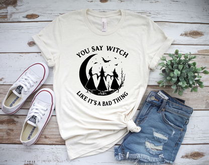 White t-shirt with witch graphic and text on a wooden surface with sneakers and shorts.