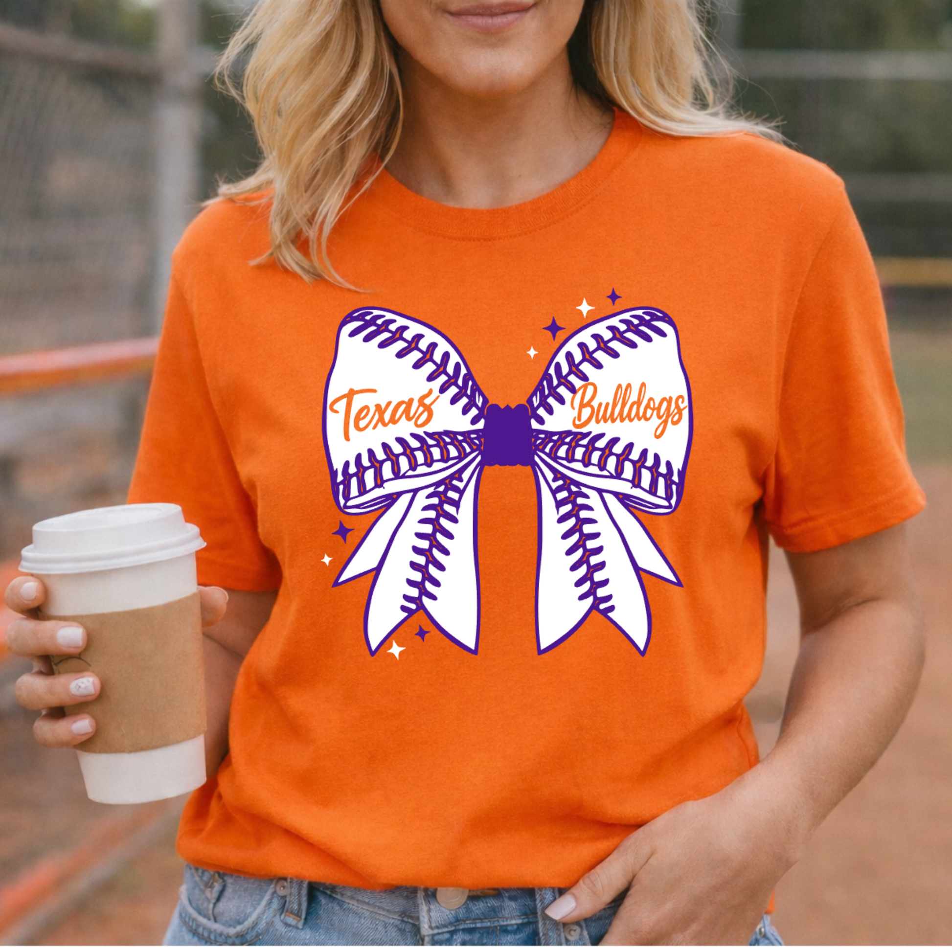 Person wearing an orange t-shirt with a bow design and 'Texas Bulldogs' text, holding a coffee cup on a sports field.