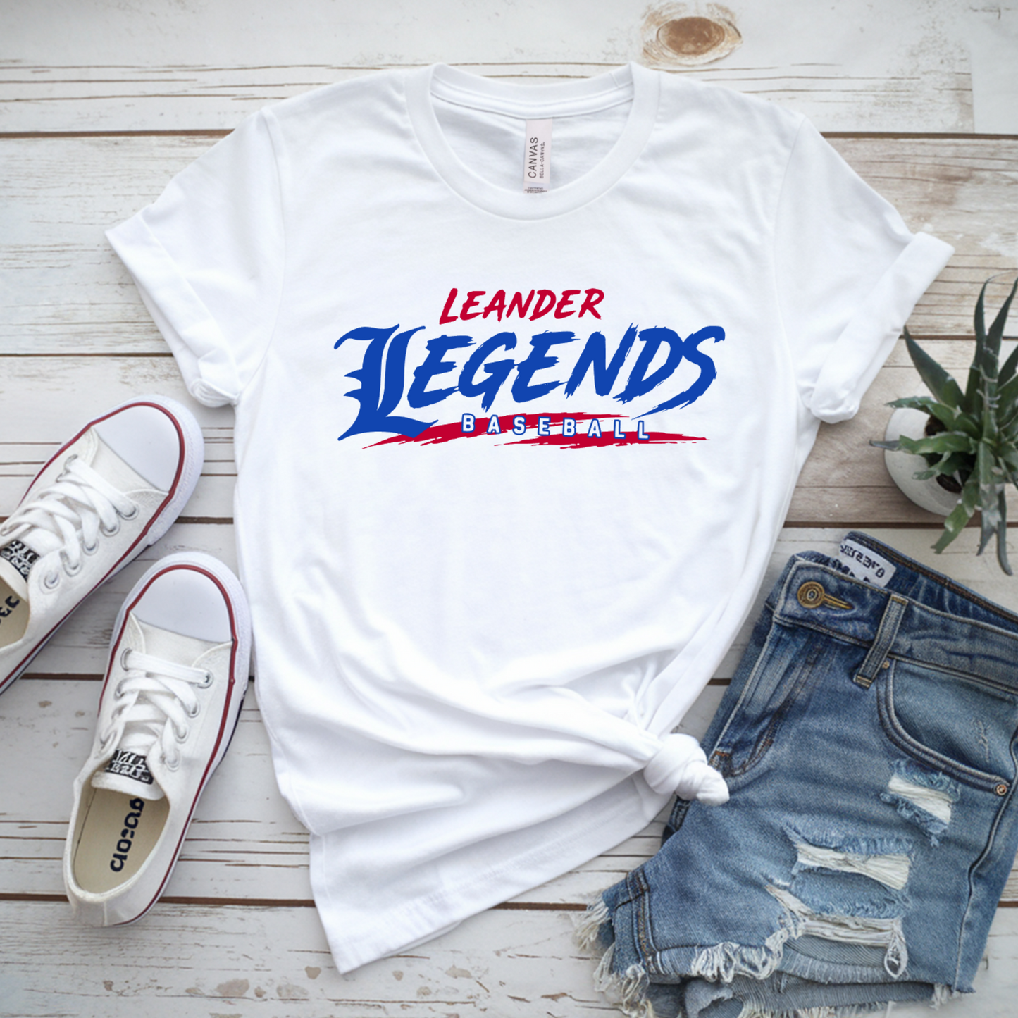 White t-shirt with 'Leander Legends' design, white sneakers, denim shorts, and a plant on a wooden surface.