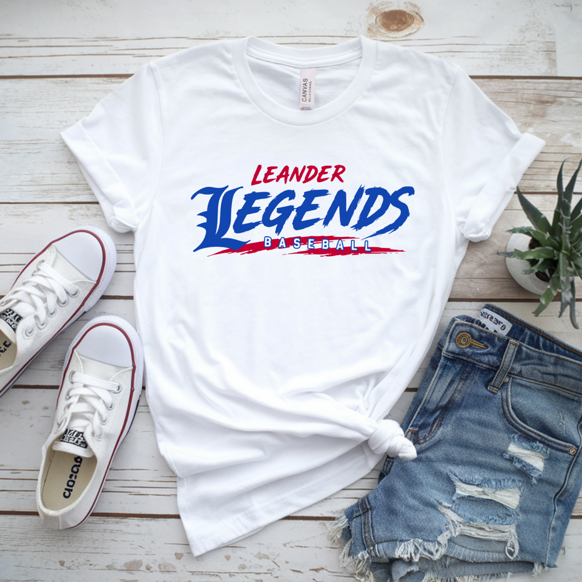 White t-shirt with 'Leander Legends' design, white sneakers, denim shorts, and a plant on a wooden surface.