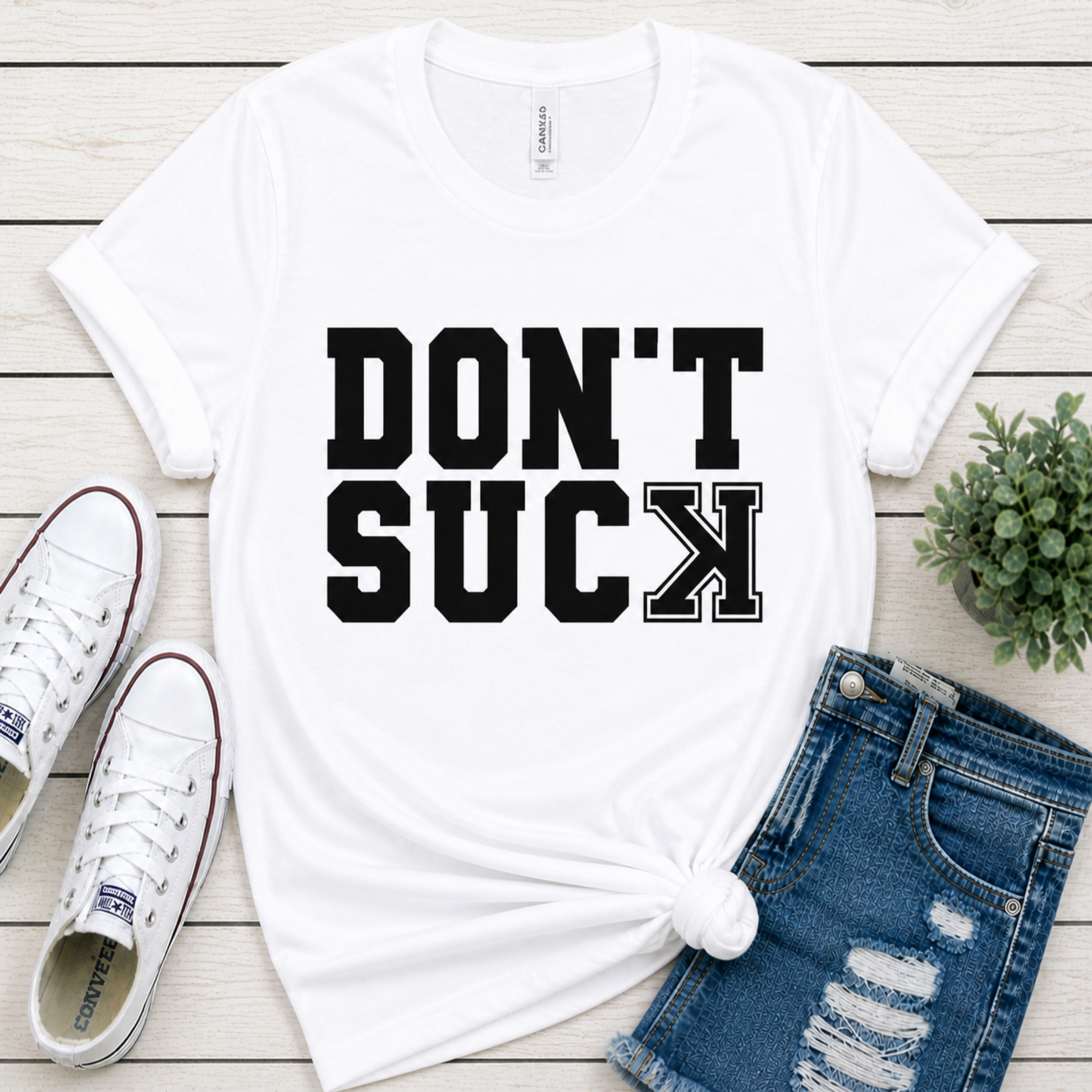 White t-shirt with 'DON'T SUCK' text, white sneakers, and denim shorts on a wooden surface.