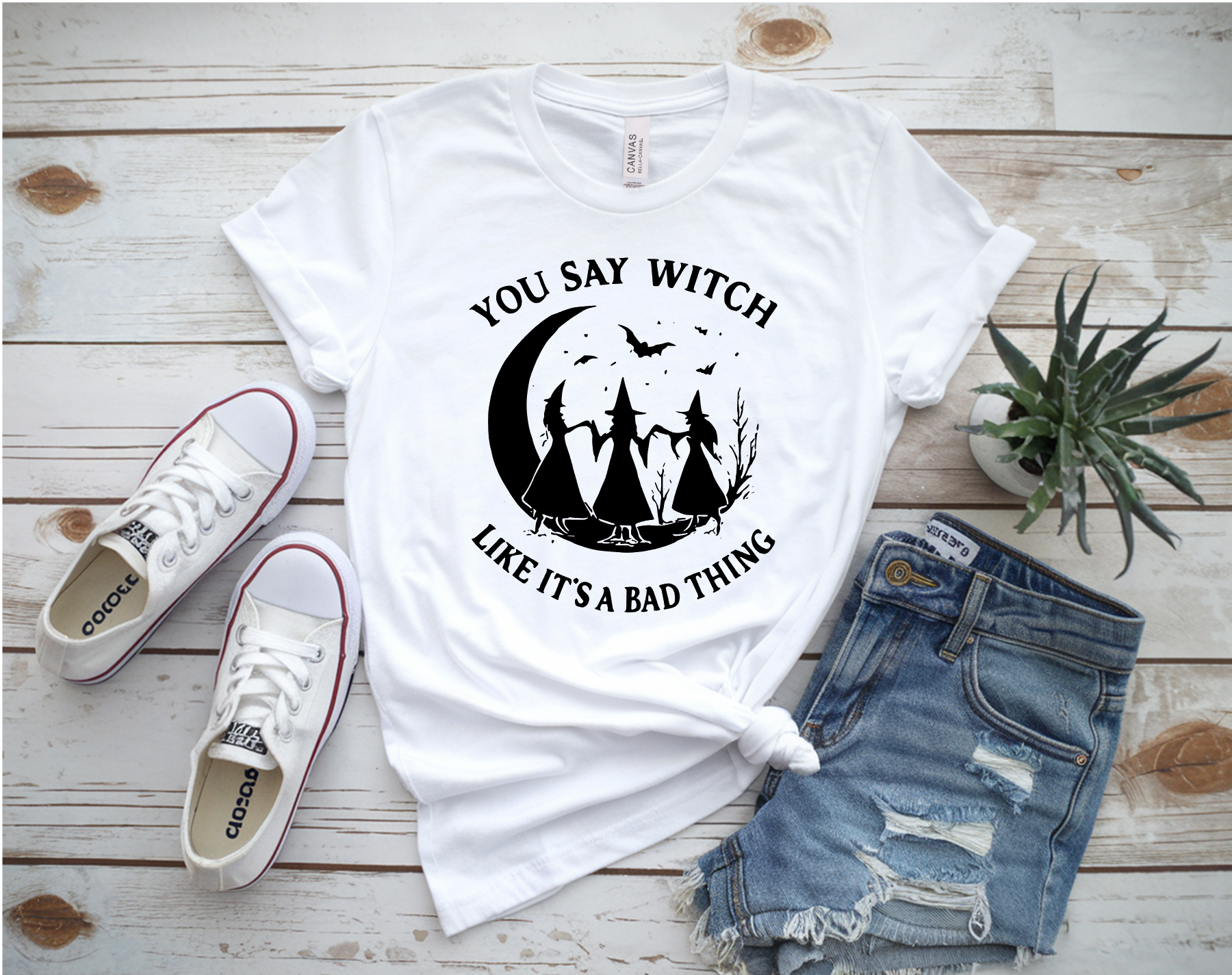 White t-shirt with 'You Say Witch Like It's a Bad Thing' design on a wooden surface with sneakers and shorts.