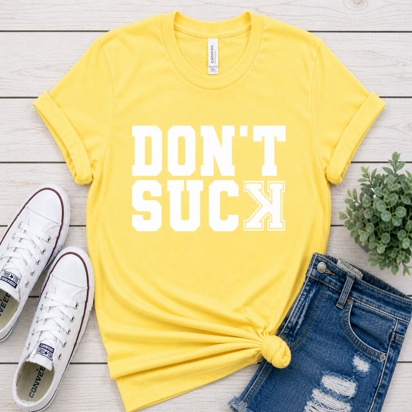 Yellow t-shirt with 'DON'T SUCK' text, white sneakers, and denim shorts on a wooden surface.